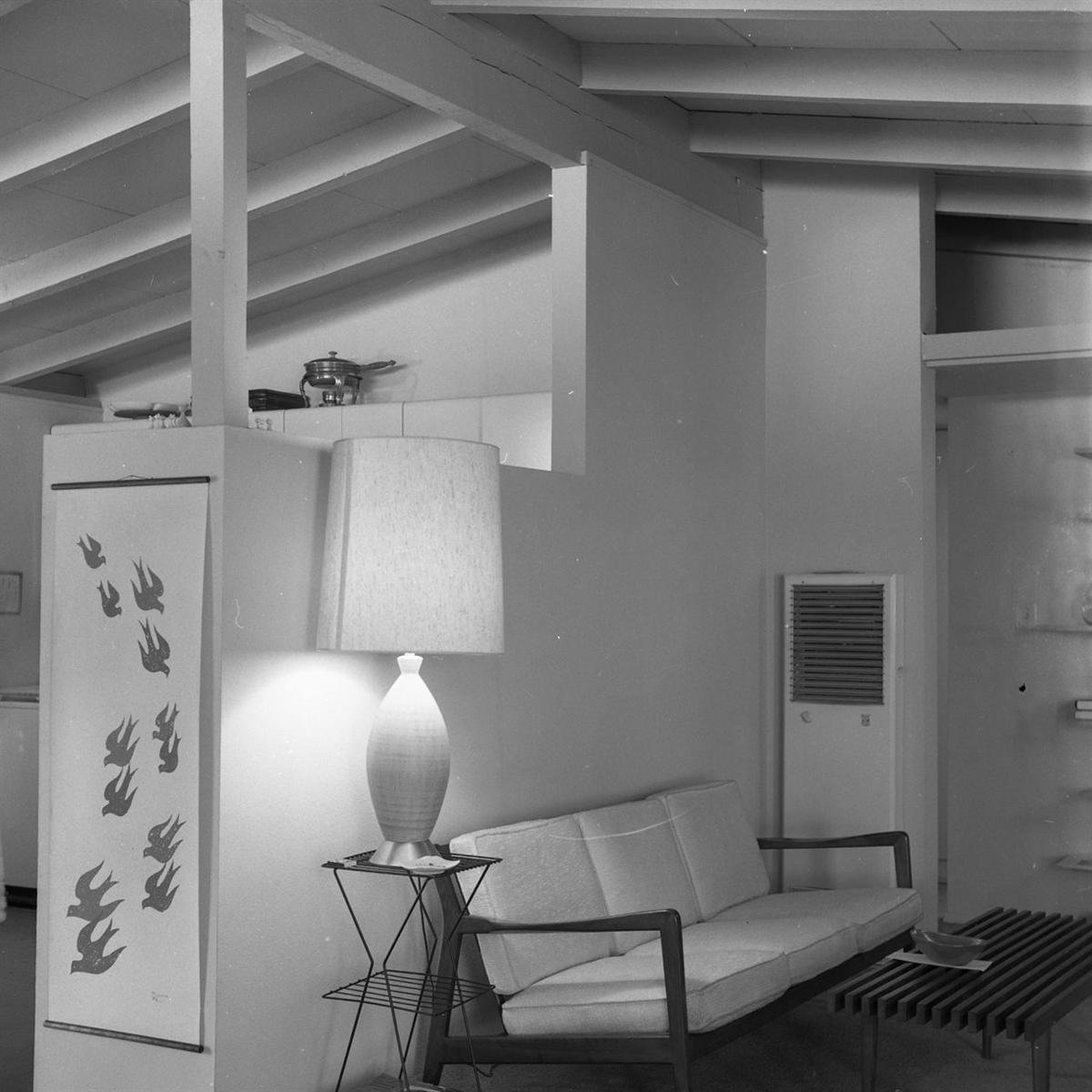 A black-and-white photograph of a 1960 California modern home interior with an open-beam ceiling, a low sofa and a ceramic lamp