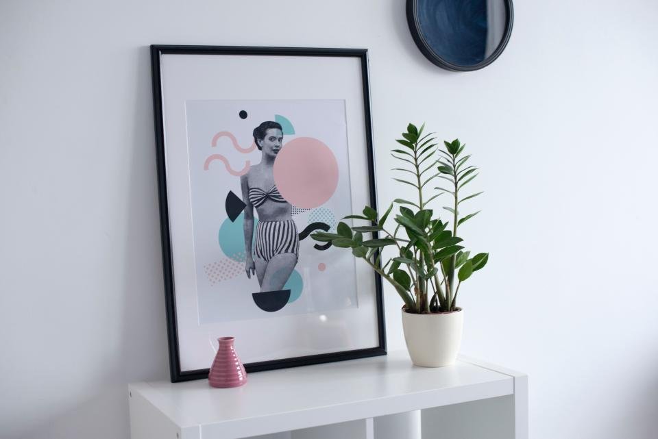 A framed graphic print leaning on a shelf beside a potted plant and a small vase