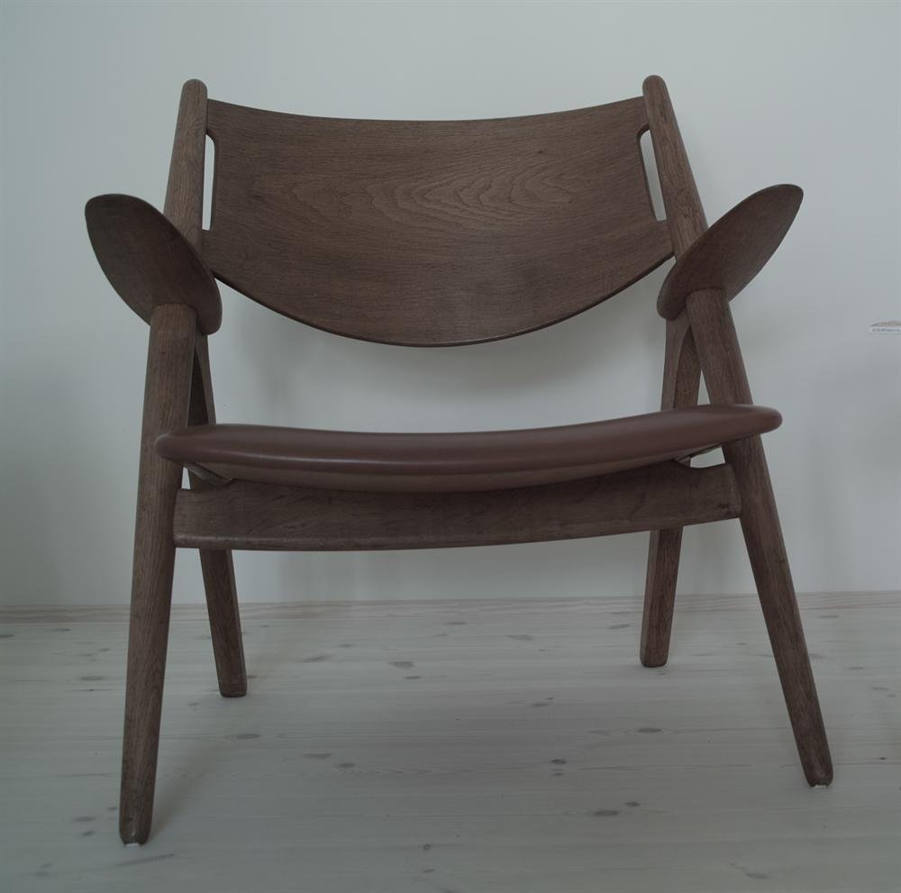 A Hans Wegner Sawhorse easy chair in oak with a leather seat, photographed head-on against a pale wall