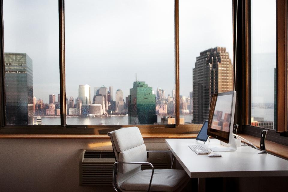 A white office chair at a desk in front of a floor-to-ceiling window with a city view